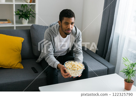 Cheerful curly haired ethnic african american male in casual clothes resting at home on comfortable sofa while watching tv channels or movie during weekend. Tv set, online television and entertiment Cheerful curly haired ethnic african american male in casual clothes resting at home on comfortable sofa while watching tv channels or movie during weekend. Tv set, online television and entertiment 122325790