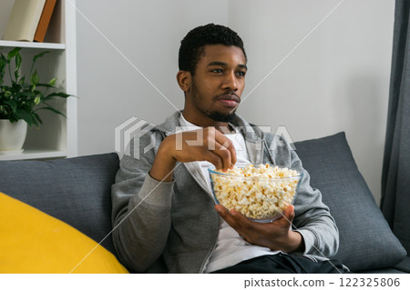 African American man sits alone on the couch and enjoying popcorn while watching television and streaming service 122325806
