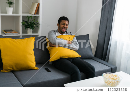 Cheerful curly haired ethnic african american male in casual clothes resting at home on comfortable sofa while watching tv channels or movie during weekend. Tv set, online television and entertiment Cheerful curly haired ethnic african american male in casual clothes resting at home on comfortable sofa while watching tv channels or movie during weekend. Tv set, online television and entertiment 122325838
