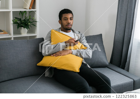 Cheerful curly haired ethnic african american male in casual clothes resting at home on comfortable sofa while watching tv channels or movie during weekend. Tv set, online television and entertiment Cheerful curly haired ethnic african american male in casual clothes resting at home on comfortable sofa while watching tv channels or movie during weekend. Tv set, online television and entertiment 122325863