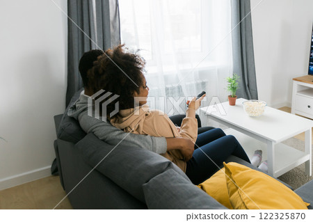 African American couple wife husband watching movie. Family pastime and leisure, watch television, rest. Relaxed man and woman watching TV series together sitting on couch at home enjoying eating African American couple wife husband watching movie. Family pastime and leisure, watch television, rest. Relaxed man and woman watching TV series together sitting on couch at home enjoying eating 122325870