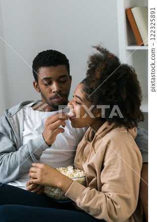 African American couple shares a playful and relaxed moment on the couch, with the man feeding popcorn to the woman. TV, series and streaming service concept African American couple shares a playful and relaxed moment on the couch, with the man feeding popcorn to the woman. TV, series and streaming service concept 122325891
