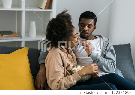 African American couple shares a playful and relaxed moment on the couch, with the man feeding popcorn to the woman. TV, series and streaming service concept African American couple shares a playful and relaxed moment on the couch, with the man feeding popcorn to the woman. TV, series and streaming service concept 122325892