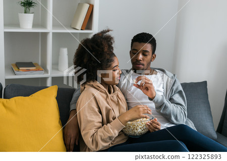 African American couple shares a playful and relaxed moment on the couch, with the man feeding popcorn to the woman. TV, series and streaming service concept African American couple shares a playful and relaxed moment on the couch, with the man feeding popcorn to the woman. TV, series and streaming service concept 122325893