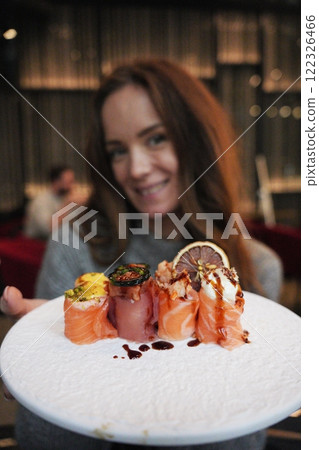 Smiling Woman Showcasing a Gourmet Sushi Platter in a Chic Restaurant Smiling Woman Showcasing a Gourmet Sushi Platter in a Chic Restaurant 122326466