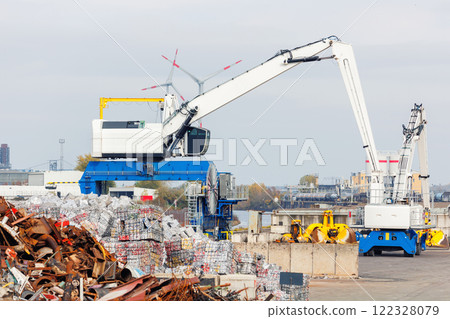 Crane at recycling facility loading large scrap metal pile with wind turbines in background. Industrial area scene waste management and resource reuse concept Crane at recycling facility loading large scrap metal pile with wind turbines in background. Industrial area scene waste management and resource reuse concept 122328079