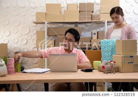 Man working at desk with laptop and various packing supplies while woman assists in organized workspace surrounded by stacked cardboard boxes 122328519