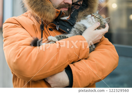 Bearded man in a fur lined hood smiles while holding a fluffy tabby cat outdoors close-up. Cozy and joyful winter scene in an urban environment, homeless animals 122329560