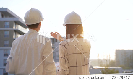 Two architects colleagues, wearing safety helmets and glasses, are examining blueprints at a construction site during sunrise or sunset, back view. Architecture and engineering Two architects colleagues, wearing safety helmets and glasses, are examining blueprints at a construction site during sunrise or sunset, back view. Architecture and engineering 122329747