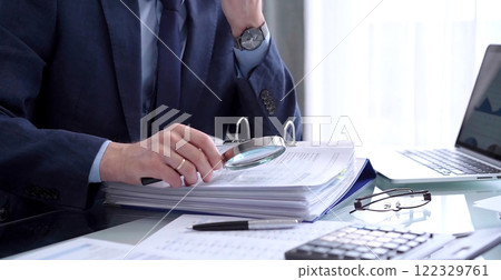 Businessman or auditor is looking through binders with papers while using a calculator and magnifying glass for financial documents analyzing. Audit and taxes concept 122329761