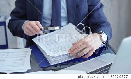 Adult businessman or auditor examining documents with magnifying glass. Professional in a suit scrutinizes financial reports, detail-focused work concept Adult businessman or auditor examining documents with magnifying glass. Professional in a suit scrutinizes financial reports, detail-focused work concept 122329777