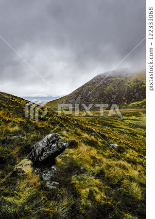 Overcast sky above a brown and green hilly terrain. Several rocks are prominent in the foreground. The scene appears somewhat barren. 122331068