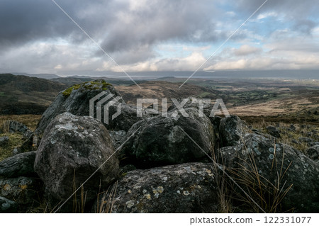 A cluster of large, dark grey rocks, lichen-covered, sits atop a grassy hill. A distant valley and cloudy sky are visible. A cluster of large, dark grey rocks, lichen-covered, sits atop a grassy hill. A distant valley and cloudy sky are visible. 122331077