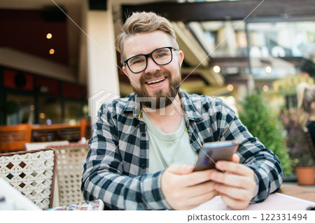 Smiling millennial man uses his smartphone while enjoying a casual outdoor cafe setting. Holidays atmosphere, modern connectivity, social networks 122331494