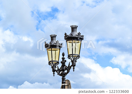 Wrought iron lamppost under a blue sky with white clouds in the Plaza des Born in Ciutadella, Menorca Wrought iron lamppost under a blue sky with white clouds in the Plaza des Born in Ciutadella, Menorca 122333600