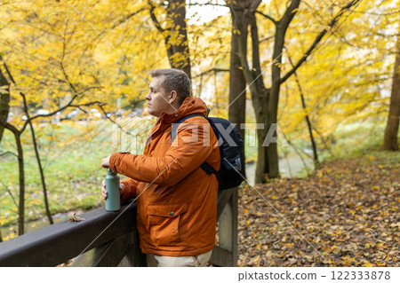 Blonde young man with thermos of tea wearing backpack standing on autumn forest trail, hiking alone. Freedom and active lifestyle concept. High quality photo 122333878