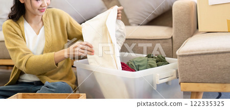 Smiling woman folding clothes for donation from a clear bin in her warm living room, promoting kindness through organization 122335325