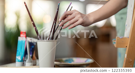 Talented artist reaching for paintbrushes in a cup while preparing for a new artwork in a bright studio 122335393