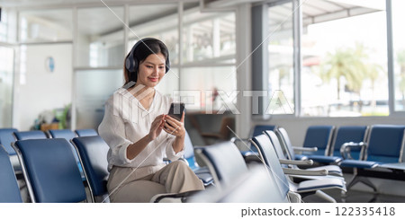 Happy young woman with headphones enjoying music while using smartphone in airport waiting area 122335418