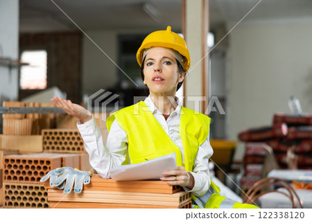 Woman foreman in a yellow vest and a protective helmet points with her hand somewhere in brick warehouse Woman foreman in a yellow vest and a protective helmet points with her hand somewhere in brick warehouse 122338120