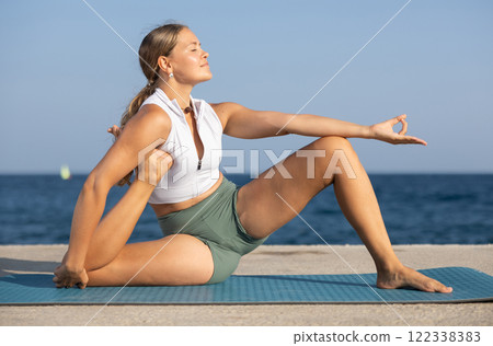 Young girl in sportwear doing stretching exercises for strong arms and abs and difficult yoga asana on the seashore on sunny day 122338383