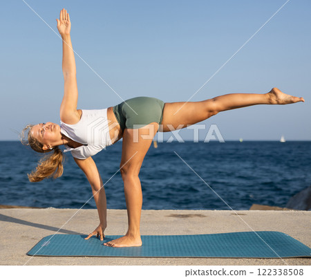On windy day young slender woman does yoga near sea doing Natarajasana 122338508