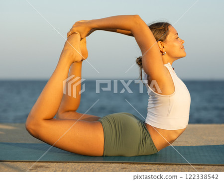 On windy day young slender woman does yoga near sea doing bow pose 122338542