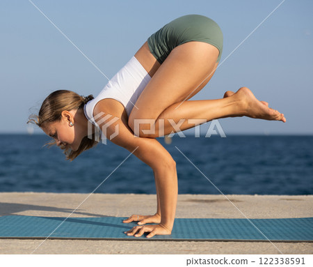 Sportive girl practicing crow pose of yoga on seashore Sportive girl practicing crow pose of yoga on seashore 122338591