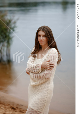 Caucasian woman in crochet beige dress standing on lake shore. Looking into camera. Caucasian woman in crochet beige dress standing on lake shore. Looking into camera. 122338733