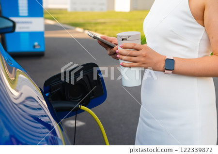 Woman in the white dresss uses her smartphone to monitor charging of an electric vehicle, holding a coffee cup. 122339872