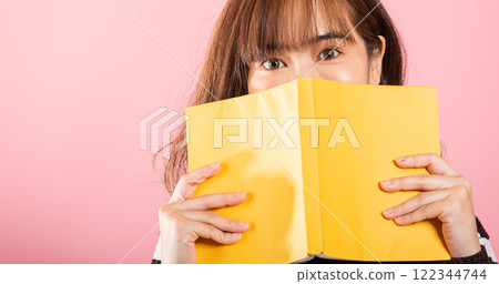 Portrait of beautiful Asian young woman teen smile covering her face with yellow book, female person hiding behind an open book show only eyes, studio shot isolated on pink background 122344744