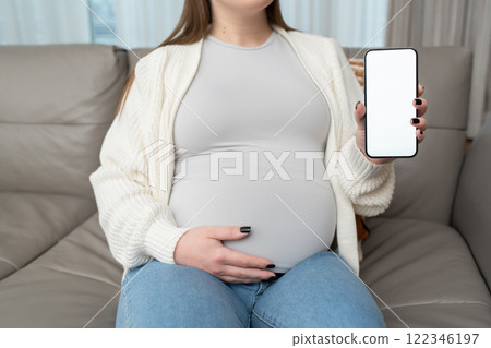 Close-up of pregnant woman holding and showing mobile phone with white empty display at home. Mockup image with copy space for app or interface customization. E-commerce. Close-up of pregnant woman holding and showing mobile phone with white empty display at home. Mockup image with copy space for app or interface customization. E-commerce. 122346197