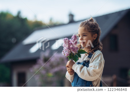 Beautiful young girl with purple lilacs flower in hands. House with solar panels in background. Beautiful young girl with purple lilacs flower in hands. House with solar panels in background. 122346848