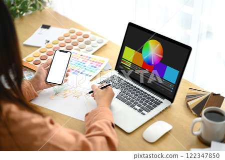 Creative woman holding smartphone working with color palette and sketches at desk 122347850