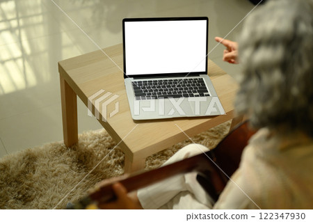 Senior woman practicing guitar in living room and following an online lesson on laptop Senior woman practicing guitar in living room and following an online lesson on laptop 122347930