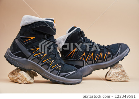 Pair of black hiking boots with orange details on small rocks. Close up of waterproof trekking shoes on beige studio background. Footwear for exploration and outdoor activities 122349581