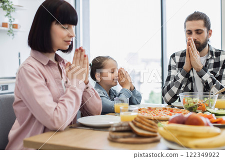 Caucasian family with parents and daughter praying together before breakfast. Meal on table includes fruit, pizza, and salad. Moment of gratitude and connection. 122349922