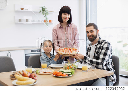 Caucasian family with parents and daughter having pizza for breakfast in bright modern kitchen. Happy child with parents sharing meal, enjoying family time and morning togetherness. 122349932
