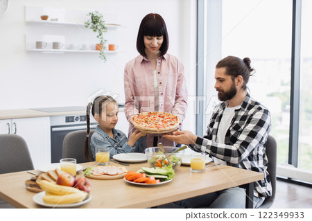 Caucasian family gathered in kitchen for breakfast with pizza, mother, father, young daughter sharing meal at table with orange juice, fruits, vegetables, cheerful atmosphere promoting family bonding. 122349933