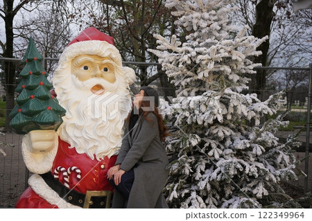 Festive woman by Santa statue and frosted Christmas tree in winter setting Festive woman by Santa statue and frosted Christmas tree in winter setting 122349964