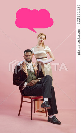 Man in baroque-inspired velvet coat sitting with elegant woman standing by his side against pink background with pink speech bubble for text. Man in baroque-inspired velvet coat sitting with elegant woman standing by his side against pink background with pink speech bubble for text. 122351185