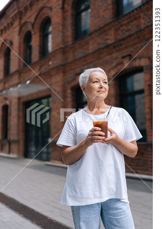 Vertical portrait of relaxed senior woman with stylish short haircut, dressed in casual attire, savoring an iced coffee while standing on city sidewalk, enjoying free time in retirement outdoors. 122351393