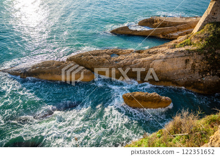 View of a sunlit rocky coastline with the sea shimmering under a clear blue sky in Ulcinj, Montenegro View of a sunlit rocky coastline with the sea shimmering under a clear blue sky in Ulcinj, Montenegro 122351652