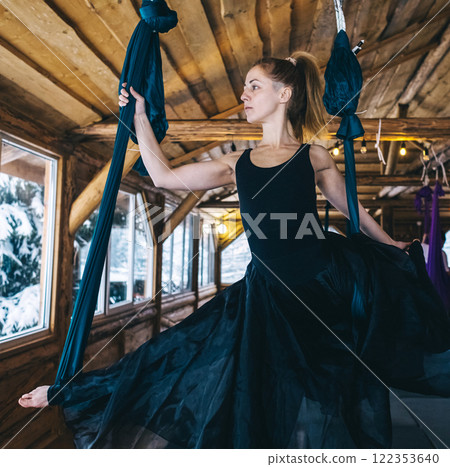 Girl gymnast in black dress doing aerial yoga. Fly yoga in hammock in spacious hall of wooden house 122353640