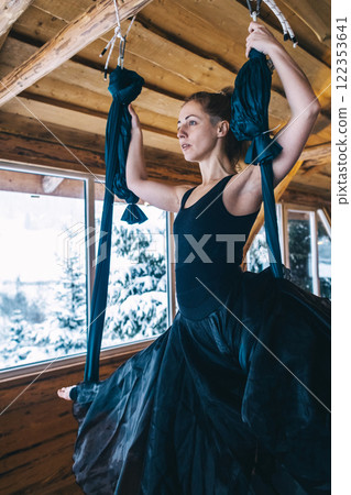 Girl gymnast in black dress doing aerial yoga. Fly yoga in hammock in spacious hall of wooden house 122353641