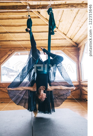 Girl gymnast in black dress doing aerial yoga. Fly yoga in hammock in spacious hall of wooden house 122353646