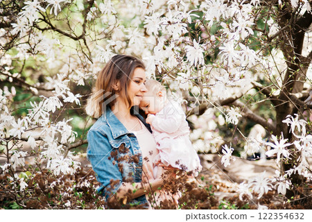 loving mother cuddling her baby in a sunny spring garden surrounded by magnolia flowers, expressing warmth and tenderness. 122354632