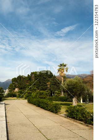 Scenic park pathway surrounded by palm trees, lush greenery, bushes, and a well-maintained lawn under an open sky Scenic park pathway surrounded by palm trees, lush greenery, bushes, and a well-maintained lawn under an open sky 122354763