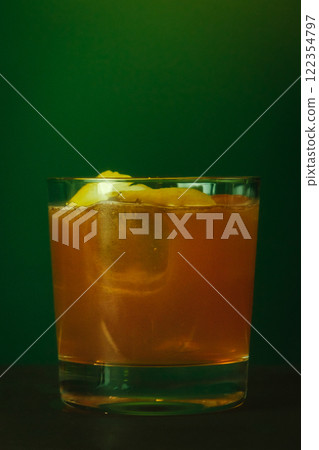 Amber Cocktail with Lemon Garnish in whiskey glass with ice cubes and citrus peel on rich green background 122354797