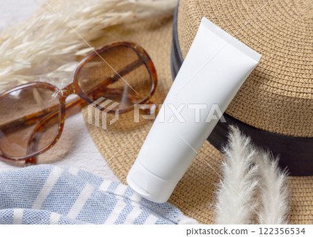 Sunscreen cream tube on hat near towel, dryed grass and sunglasses closeup, cosmetic mockup 122356534